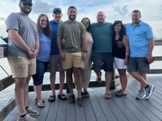 Group of seven friends standing on a dock by the water on a sunny day.