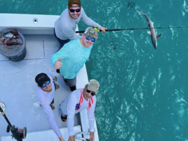 Four people on a boat holding a fishing rod with a caught fish above turquoise water.